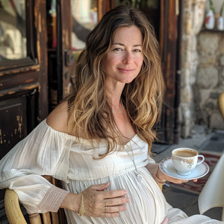Femme enceinte âgée avec une expression douce, habillée avec une robe blanche, elle est à la terrasse d'un café et a une tasse da café dans la main