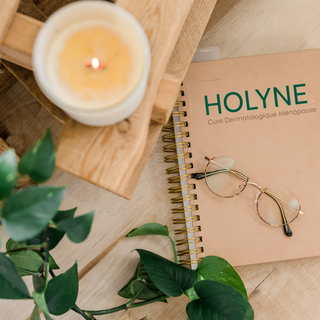 Un moment de détente HOLYNE avec une bougie allumée, des lunettes posées sur un carnet indiquant 'Cure Dermatologique Ménopause', évoquant un bien-être holistique pour la peau des femmes Ménopauseés
