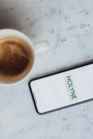 Smartphone affichant la page d'accueil de HOLYNE sur un comptoir en marbre à côté d'une tasse de café, évoquant une pause bien-être dans le quotidien.